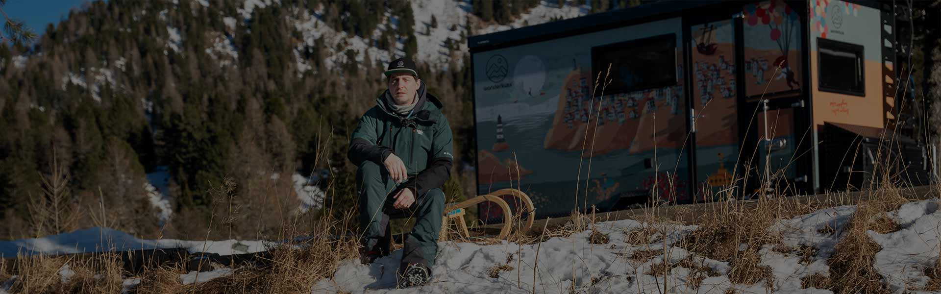 Wintercamping Packliste - Wanderkuss Sportwohnwagen im Hintergrund, davor sitzt Christoph auf der Rodel im Schnee